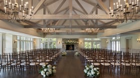 Indoor wedding ceremony setup at The Barker House in Scituate, Massachusetts, featuring wooden beams, chandeliers, and a stone fireplace adorned with white floral arrangements.