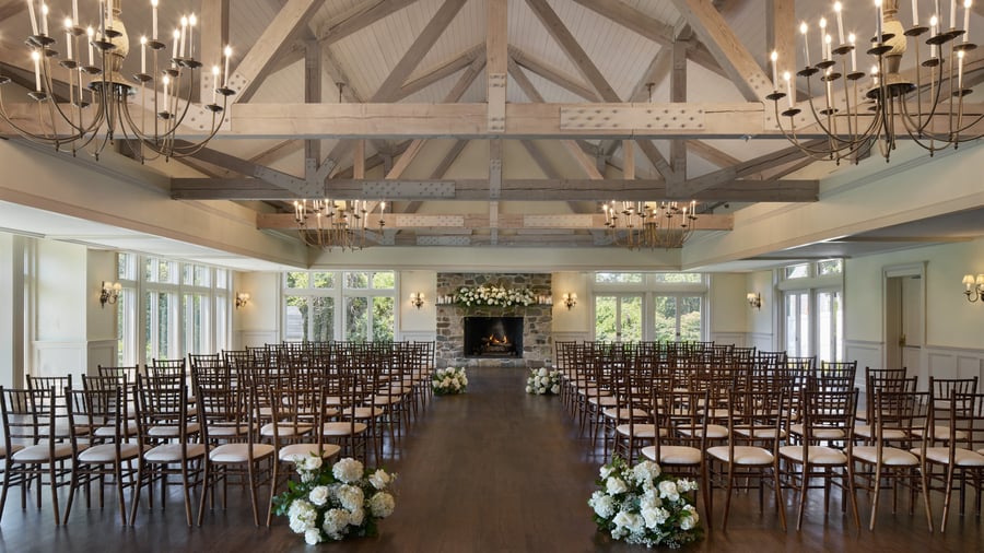 Indoor wedding ceremony setup at The Barker House in Scituate, Massachusetts, featuring wooden beams, chandeliers, and a stone fireplace adorned with white floral arrangements.