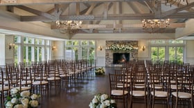 Elegant indoor ceremony setup at The Barker House featuring rows of wooden chairs facing a stone fireplace decorated with lush white flowers and candles.