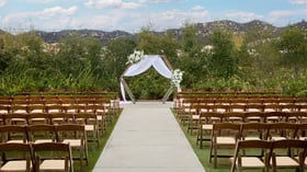 Wooden chairs lined along an outdoor ceremony aisle leading to a hexagon arch draped in white fabric and florals at Canopy Grove wedding venue in Escondido, California.