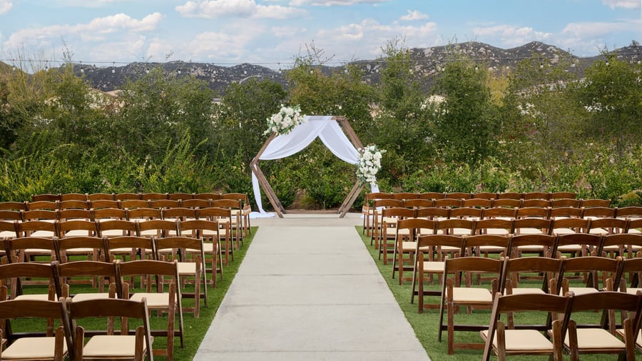 Wooden chairs lined along an outdoor ceremony aisle leading to a hexagon arch draped in white fabric and florals at Canopy Grove wedding venue in Escondido, California.