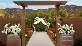 Wooden pergola entrance with wine barrels topped with flowers leading to an outdoor ceremony aisle and arch at Canopy Grove wedding venue.