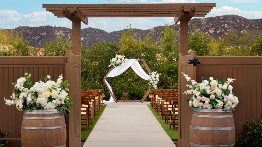 Wooden pergola entrance with wine barrels topped with flowers leading to an outdoor ceremony aisle and arch at Canopy Grove wedding venue.