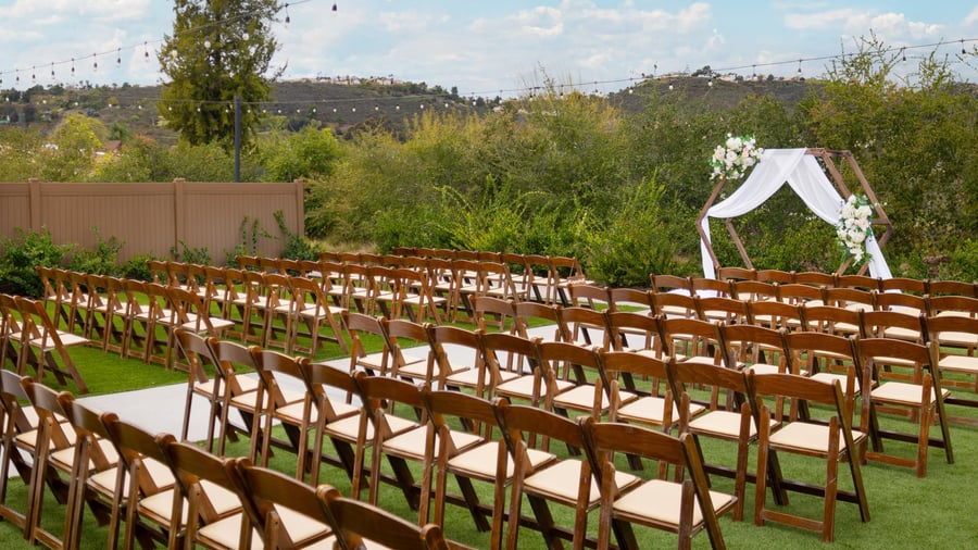 Side view of rows of wooden chairs facing a decorated ceremony arch on the outdoor lawn at Canopy Grove in Escondido, CA.