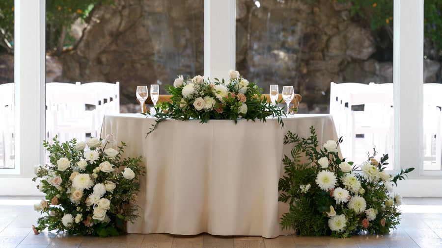 Elegant sweetheart table at Colby Falls Grand Hall in Arizona with lush floral arrangements and champagne flutes by floor-to-ceiling windows.