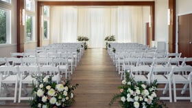 Indoor wedding ceremony space at Colby Falls in Arizona with white drapery backdrop, wooden floors, and rows of white chairs with floral