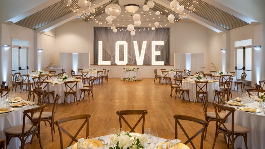 Sophisticated indoor wedding reception at Cuvier Club by Wedgewood Weddings featuring illuminated LOVE marquee sign, cross-back wooden chairs, white linens, paper lantern ceiling, and hardwood floors in La Jolla