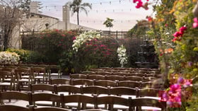 Beautiful Garden Courtyard wedding ceremony setup at Cuvier Club La Jolla with wooden chairs, white floral ceremony arch, colorful bougainvillea, and fountain backdrop