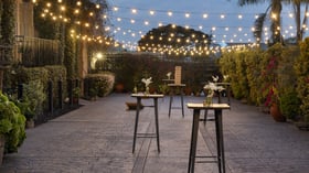 Romantic evening cocktail hour in the Garden Courtyard at Cuvier Club La Jolla with string bistro lights, high-top tables, white floral arrangements, and tropical landscaping