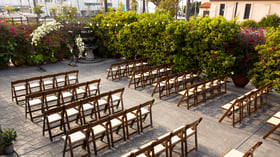 Intimate outdoor wedding ceremony space in the Garden Courtyard at Cuvier Club featuring wooden cross-back chairs, white floral arch, fountain backdrop, and vibrant bougainvillea blooms