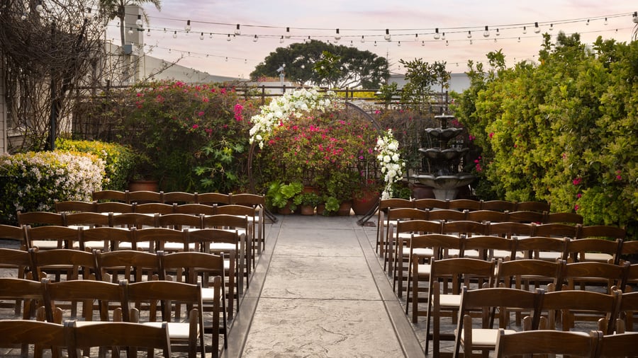 Elegant outdoor wedding ceremony setup in the Garden Courtyard at Cuvier Club by Wedgewood Weddings in La Jolla, featuring wooden chairs arranged for guests with lush bougainvillea blooms and string lights overhead