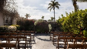 Beautiful outdoor wedding ceremony in the Garden Courtyard at Cuvier Club featuring white floral arch, wooden cross-back chairs, string lights, and tropical bougainvillea in La Jolla
