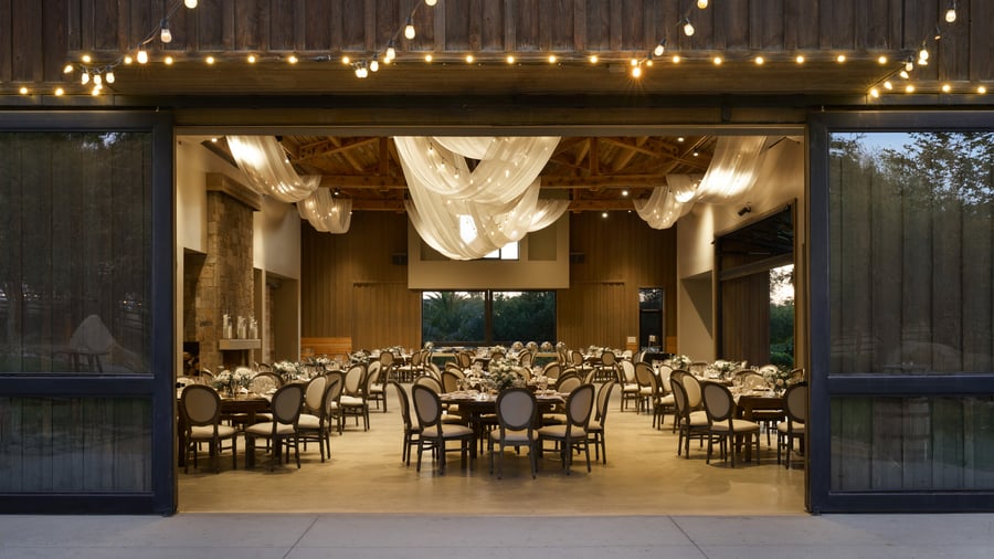Elegant evening reception at Galway Downs viewed through open barn doors, showcasing draped ceiling lighting, round tables with neutral linens, and warm interior ambiance perfect for Temecula weddings