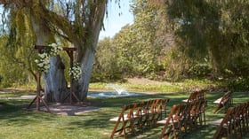 Beautiful lakeside wedding ceremony at The Meadows, Galway Downs featuring rustic wooden arch with white floral arrangements, wooden folding chairs, and serene water feature surrounded by mature trees in Temecula, California
