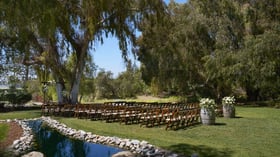 Romantic lakeside wedding ceremony at The Meadows, Galway Downs featuring rows of wooden chairs, wine barrel floral arrangements, and a stunning natural water backdrop surrounded by willow trees in Temecula