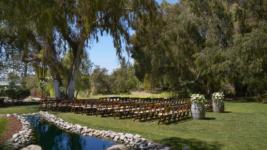 Romantic lakeside wedding ceremony at The Meadows, Galway Downs featuring rows of wooden chairs, wine barrel floral arrangements, and a stunning natural water backdrop surrounded by willow trees in Temecula