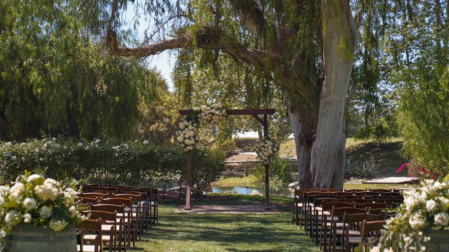 Elegant outdoor wedding ceremony setup at The Meadows, Galway Downs featuring rustic wooden chairs, white floral arrangements, and a romantic arch under majestic willow trees in Temecula, California