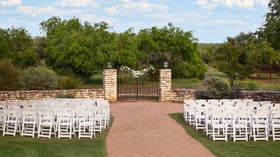 White chairs arranged for a wedding ceremony facing the iconic stone and iron gates at Hofmann Ranch in Castroville, TX.