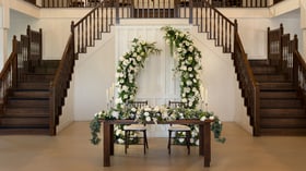 Romantic sweetheart table in the Grande Hall at Hofmann Ranch featuring white floral arch, lush greenery, and classic wooden staircase backdrop.