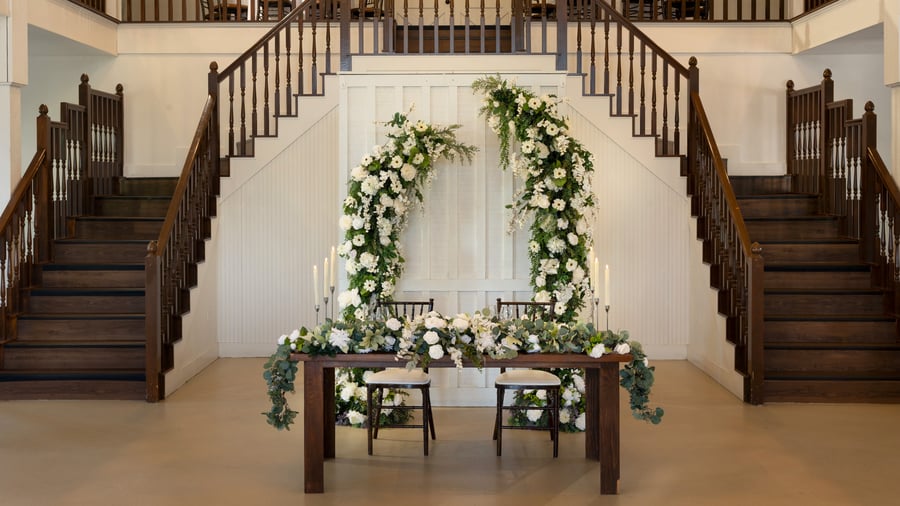 Romantic sweetheart table in the Grande Hall at Hofmann Ranch featuring white floral arch, lush greenery, and classic wooden staircase backdrop.