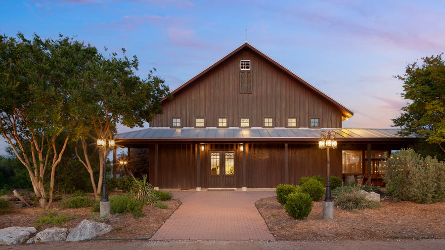 Evening view of Hofmann Ranch in Castroville, TX, showcasing its rustic barn-style exterior, warm lighting, and landscaped entrance surrounded by Texas greenery.