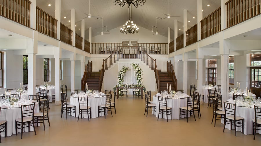 The Grande Hall at Hofmann Ranch set for a wedding reception with round tables, white linens, floral centerpieces, and dramatic dual staircases as the backdrop.