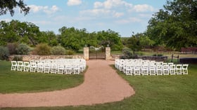 Outdoor wedding ceremony at Hofmann Ranch in Castroville, TX, with white chairs facing a rustic stone and iron gate altar framed by Texas hill country scenery.