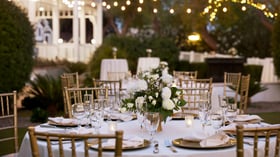 Close-up of elegant wedding reception table at Lindsay Grove in Mesa, AZ with white linens, floral centerpiece, gold chiavari chairs, and twinkle lights overhead.