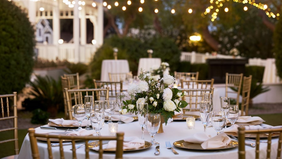 Close-up of elegant wedding reception table at Lindsay Grove in Mesa, AZ with white linens, floral centerpiece, gold chiavari chairs, and twinkle lights overhead.