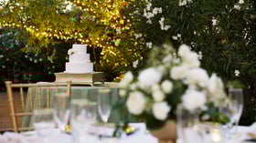 White tiered wedding cake on gold stand at Lindsay Grove in Mesa, AZ with greenery, twinkle lights, and floral backdrop.
