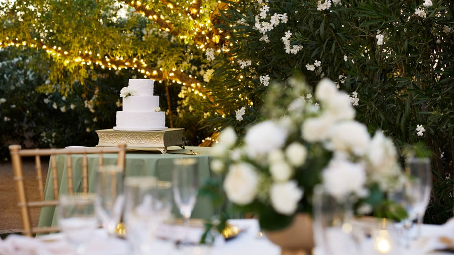 White tiered wedding cake on gold stand at Lindsay Grove in Mesa, AZ with greenery, twinkle lights, and floral backdrop.