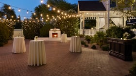 Outdoor cocktail hour setup at Lindsay Grove in Mesa, AZ featuring tall cocktail tables with white linens, glowing string lights, and a cozy fireplace.