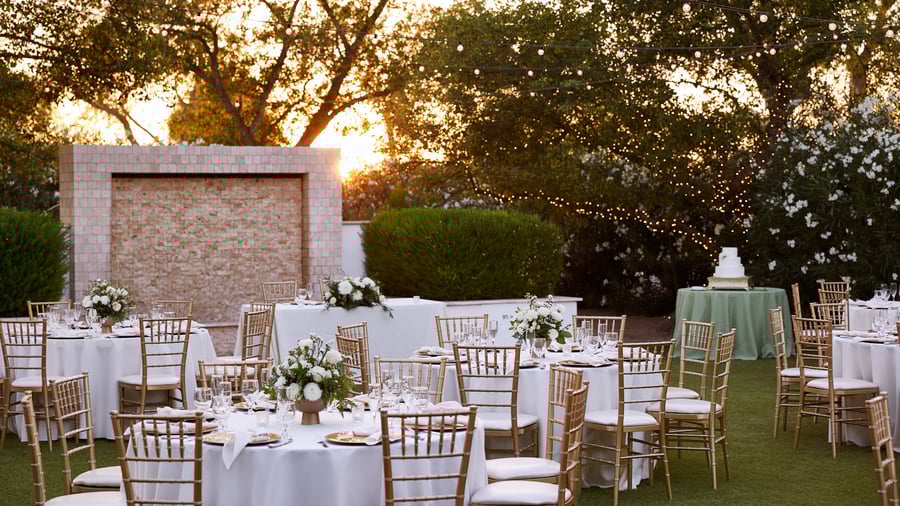 Evening reception at Lindsay Grove in Mesa, AZ with white table linens, gold chiavari chairs, floral centerpieces, and string lights against a sunset backdrop.