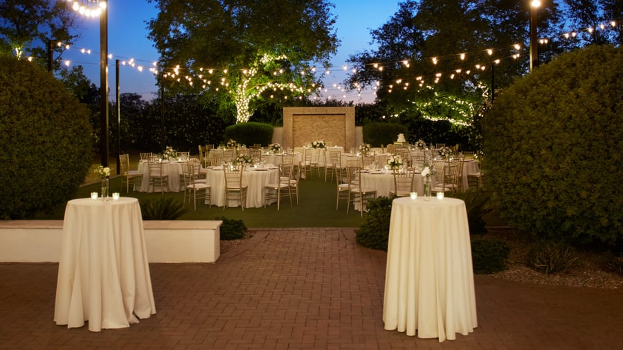 Brick patio cocktail reception at Lindsay Grove in Mesa, AZ with high-top tables, floral accents, and twinkle lights illuminating the garden.
