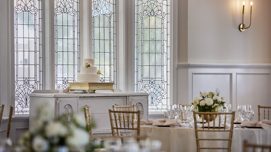 Elegant tiered wedding cake on a gold stand at Lindsay Grove Grand Hall in Mesa, AZ with stained glass windows and floral table décor.