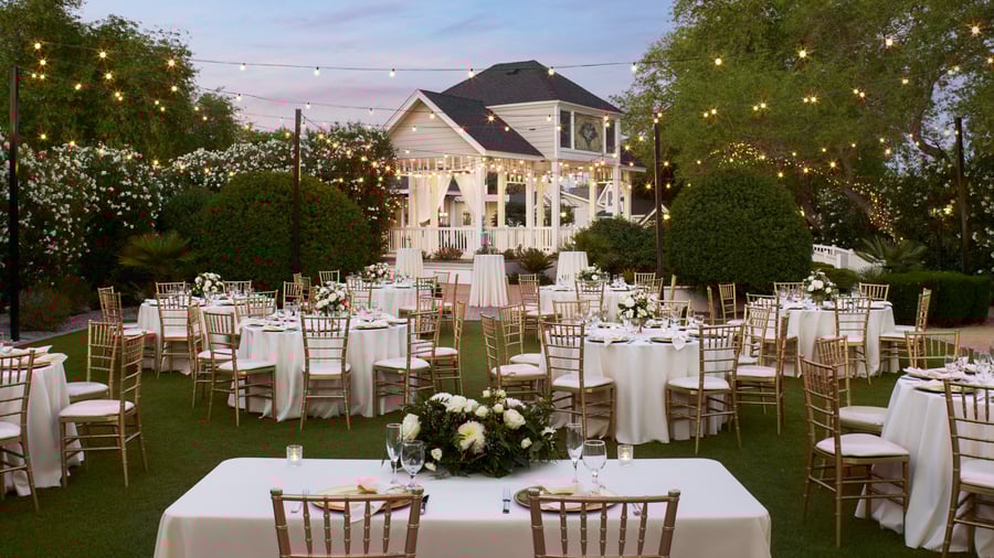 Romantic outdoor wedding reception on the brick patio at Lindsay Grove in Mesa, AZ featuring tables with white linens, gold chairs, and glowing string lights.