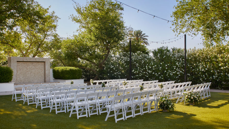 Scenic outdoor ceremony setup at Lindsay Grove in Mesa, AZ with rows of white chairs, floral arrangements, and a stone wall backdrop.