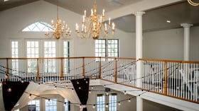Elegant two-story ballroom interior featuring ornate brass chandeliers, arched windows, wooden balcony railings, and decorative string lights with draped fabric bunting at Lionscrest Manor wedding venue in Lyons, Colorado