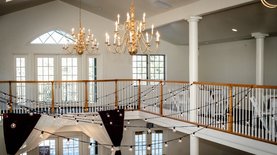 Elegant two-story ballroom interior featuring ornate brass chandeliers, arched windows, wooden balcony railings, and decorative string lights with draped fabric bunting at Lionscrest Manor wedding venue in Lyons, Colorado