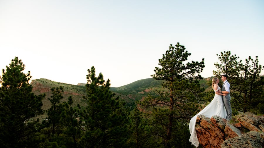 Bride in flowing white gown with dramatic train cascading over red rock cliff and groom in grey vest embracing on scenic overlook with sweeping Colorado mountain and pine forest views near Lionscrest Manor in Lyons