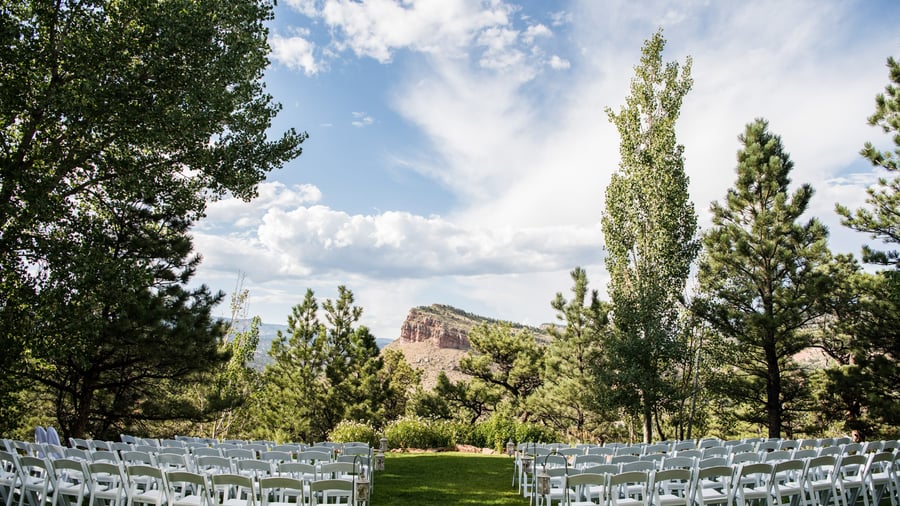 Outdoor wedding ceremony with white chairs arranged on green lawn overlooking dramatic Colorado mountain formations framed by pine and aspen trees at Lionscrest Manor in Lyons