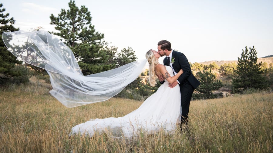 Bride in white ballgown with dramatically windswept cathedral veil and groom in black suit kissing in golden grass meadow with pine trees and Colorado foothills near Lionscrest Manor in Lyons