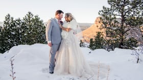 Bride in champagne ballgown with white fur wrap and groom in grey suit embracing in fresh snow with snow-covered pine trees and iconic Colorado red rock mesa backdrop at Lionscrest Manor winter wedding