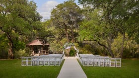 Elegant outdoor wedding ceremony setup at Scenic Springs in Helotes, Texas, with white chairs, a floral arch, and a tree-lined forest backdrop.
