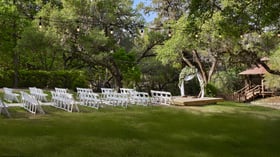 Outdoor wedding ceremony setup at Scenic Springs in Helotes, Texas, with floral arch, gazebo, and white seating under oak trees.