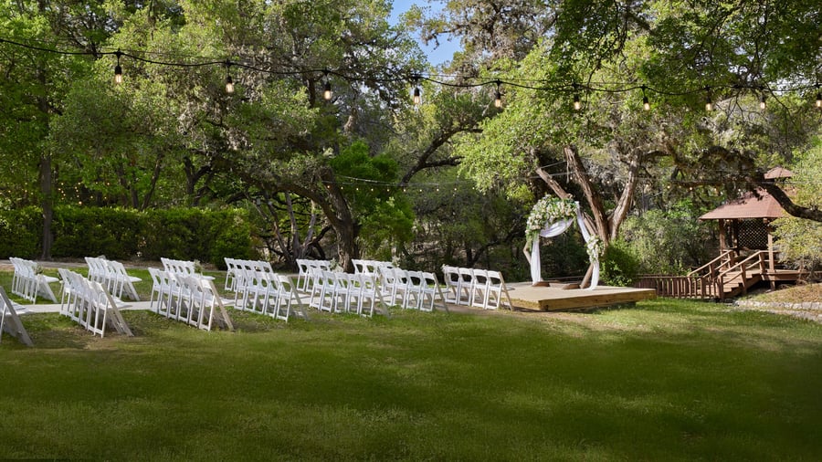 Outdoor wedding ceremony setup at Scenic Springs in Helotes, Texas, with floral arch, gazebo, and white seating under oak trees.