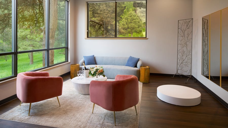 Stylish wedding suite at The Pines featuring blush velvet chairs, a soft blue sofa, large windows with forest views, and a white floral arrangement on a coffee table.
