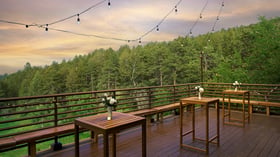 Scenic outdoor deck at The Pines featuring wooden cocktail tables, floral vases, and string lights overlooking lush green forest hills at sunset.