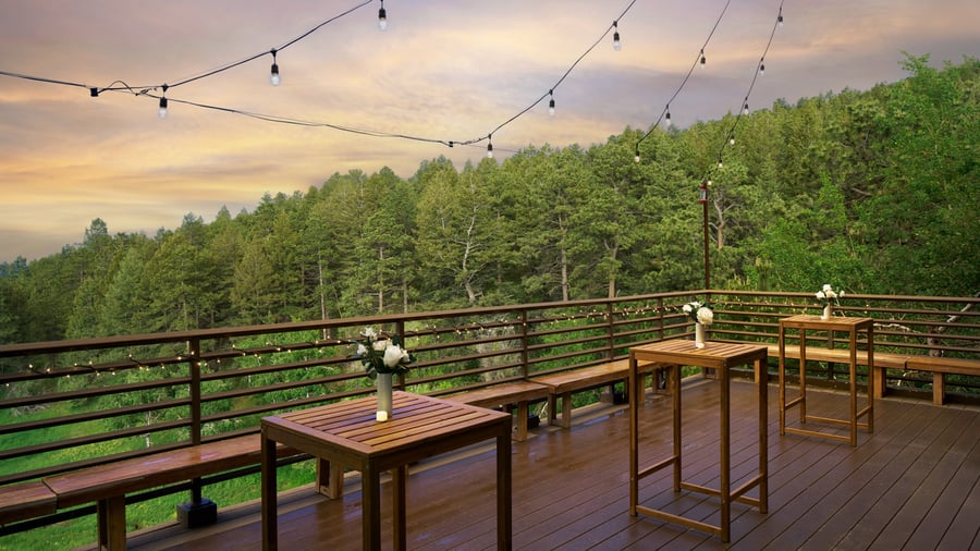 Scenic outdoor deck at The Pines featuring wooden cocktail tables, floral vases, and string lights overlooking lush green forest hills at sunset.