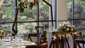 Sweetheart table at The Pines adorned with white and blush florals, rustic wooden chairs, and a geometric floral arch beside large windows overlooking forest scenery.